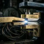 A close-up of metal being welded with bright sparks in a dark industrial setting, featuring gold and grey tones