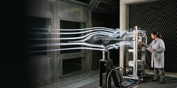 A dark wind tunnel test reveals airflow around a dark, streamlined model, with a scientist monitoring equipment nearby.