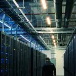 Long data centre aisle between dark server racks under cable trays, a figure walking away
