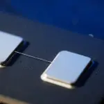 A close-up of a dark circuit board with two metallic squares connected by a thin wire, illuminated with cool blue lighting.
