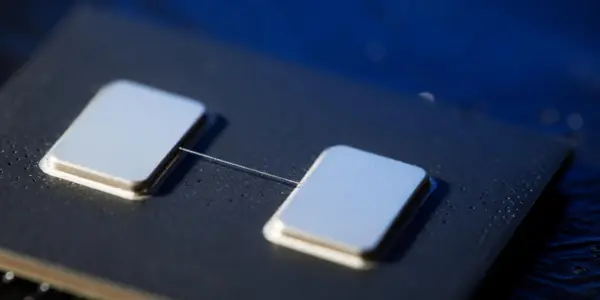 A close-up of a dark circuit board with two metallic squares connected by a thin wire, illuminated with cool blue lighting.