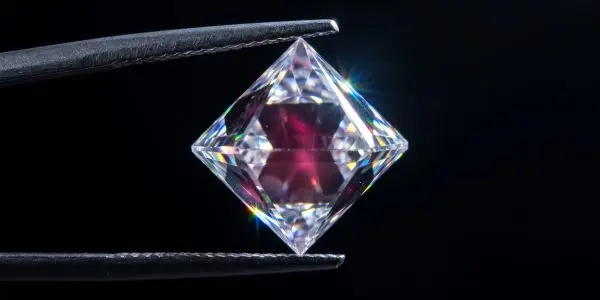 Clear faceted diamond with a pink inner glow held between metal tweezers on black