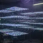 A complex, multi-layered structure of illuminated planes and connecting lines fills a darkened laboratory space, dominated by blue and purple hues.