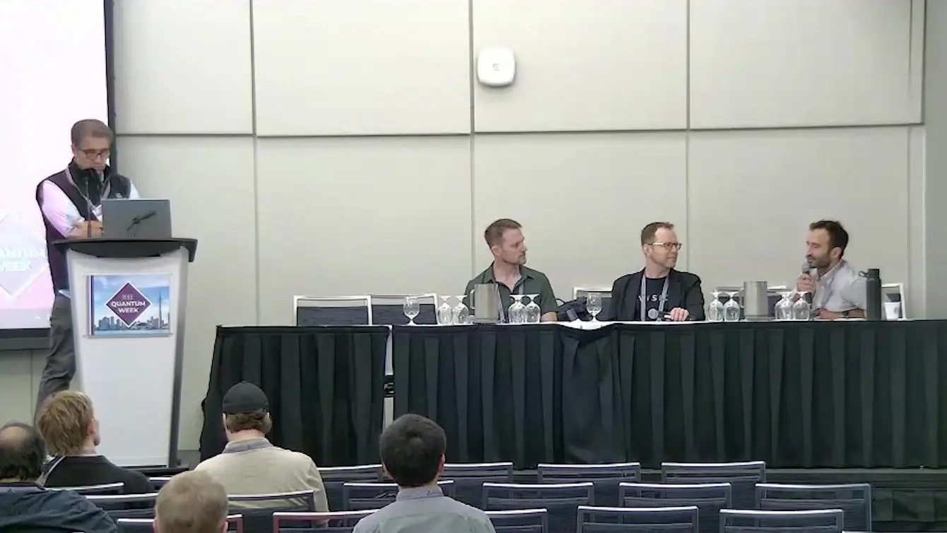 Panel on spin qubits linked by light at IEEE Quantum Week 2026, with a chair at the podium and three guests.