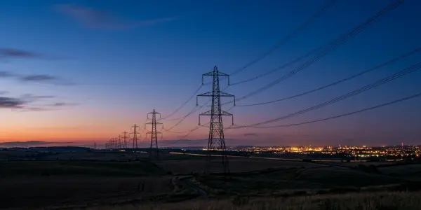 A line of transmission pylons crossing open country at dusk with city lights on the horizon
