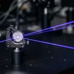 Blue laser beams cross at a small mirror mount on a dark optical table