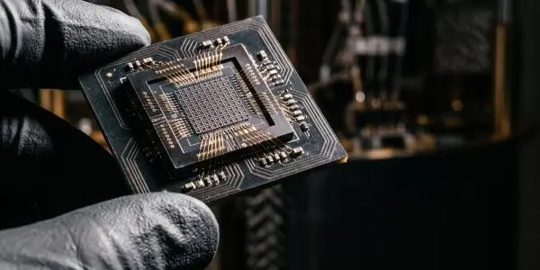 A gloved hand holds a quantum processor chip with a gridded core and fine gold wire bonds