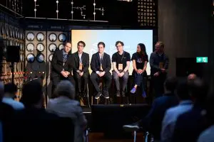 A panel discussion takes place on a stage with a backdrop of electrical diagrams, in shades of grey and black.