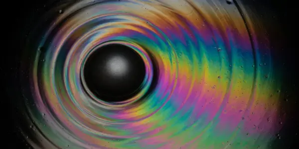 A swirling, iridescent vortex of colour surrounds a dark central sphere against a black background, creating an abstract, otherworldly effect.