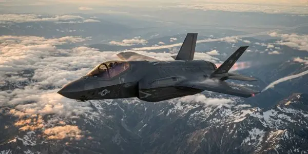 Illustration of a stealth fighter jet with US markings flying above snow-capped mountains and clouds at sunset