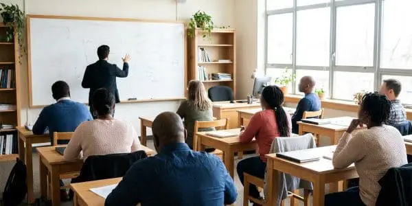 Adult learners at desks facing a whiteboard of faint diagrams