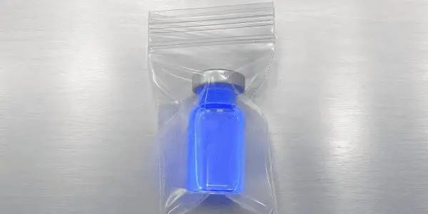 A small blue vial containing a liquid is encased in a clear plastic bag on a reflective, metallic surface.