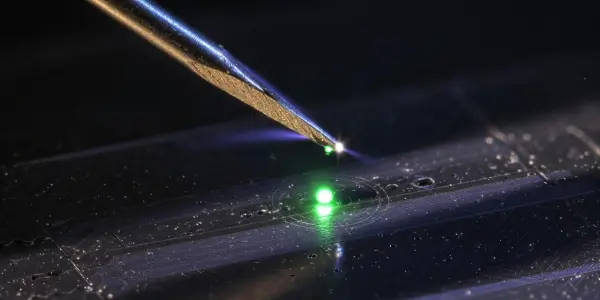 A metallic probe touches a circuit board with a green laser spot, set against a dark background with blue highlights.