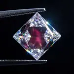 Clear faceted diamond with a pink inner glow held between metal tweezers on black