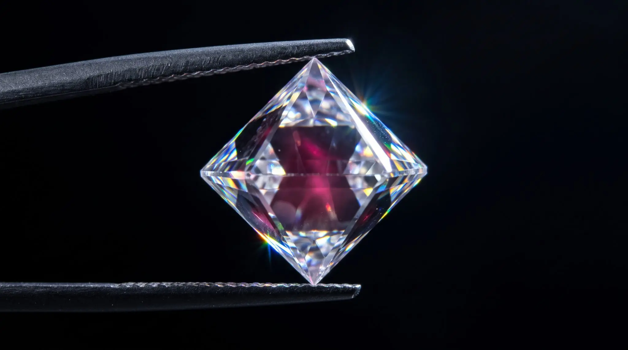 Clear faceted diamond with a pink inner glow held between metal tweezers on black