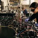A scientist works with complex optical equipment in a laboratory filled with wires and glowing beams, dominated by silver, blue and black.