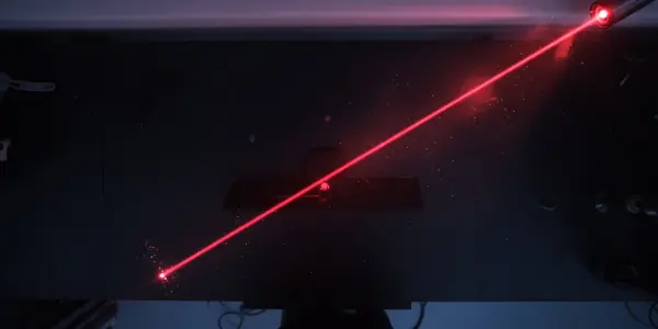 A bright red laser beam cuts through a dark laboratory, illuminating dust particles and reflecting off metallic surfaces.