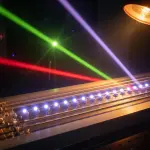 A dark laboratory setting features a complex metallic apparatus illuminated by red, green, and purple laser beams and a lamp.