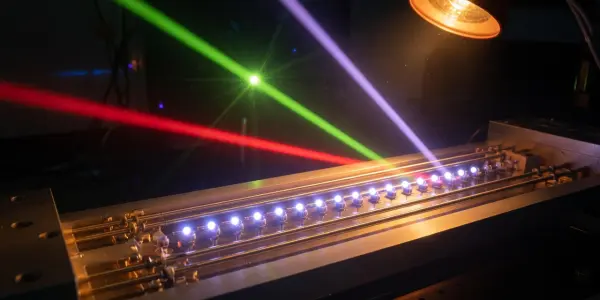 A dark laboratory setting features a complex metallic apparatus illuminated by red, green, and purple laser beams and a lamp.