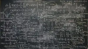 A chalkboard covered in complex quantum physics equations dominates the frame, set in a dimly lit room.