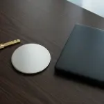 A dark wood surface holds a brass key, a circular device, and a closed laptop, lit by window light.