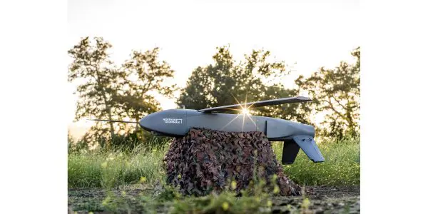 A grey unmanned aerial vehicle rests on a camouflage blanket in a grassy field with trees in the background.