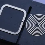 A dark, moody macro shot features two metallic micro-fabricated shapes—a square and a spiral—etched onto a black substrate.