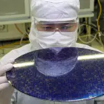A person in a cleanroom holds a circular wafer with intricate blue and silver circuitry, illuminated by bright overhead lights.