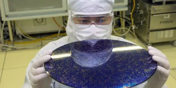 A person in a cleanroom holds a circular wafer with intricate blue and silver circuitry, illuminated by bright overhead lights.