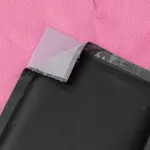 A partially opened pink padded envelope reveals a black, silver-edged rectangular package, set against a bright pink background.