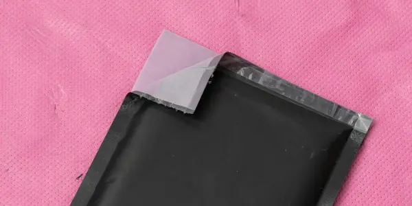 A partially opened pink padded envelope reveals a black, silver-edged rectangular package, set against a bright pink background.