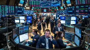 A bustling stock exchange floor displays numerous computer screens filled with financial data in red, green, and grey.