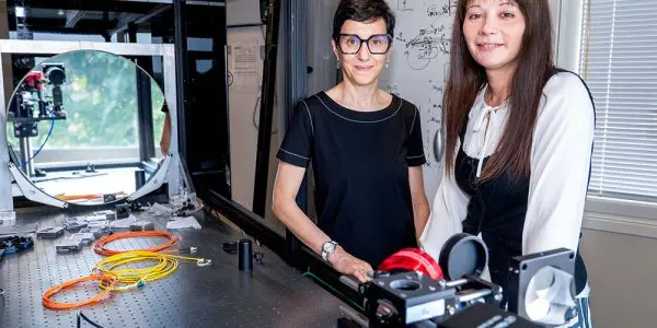Two scientists stand beside complex optical equipment in a dark laboratory setting with grey, white, and red elements.