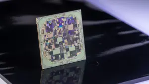 A square microchip with intricate circuitry sits on a reflective black surface, illuminated by bright, directional lighting.