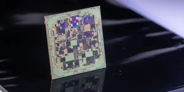 A square microchip with intricate circuitry sits on a reflective black surface, illuminated by bright, directional lighting.