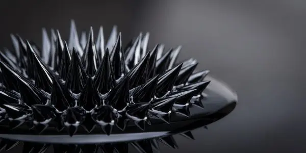 Glossy black ferrofluid rising into sharp spikes on a dish against a grey backdrop