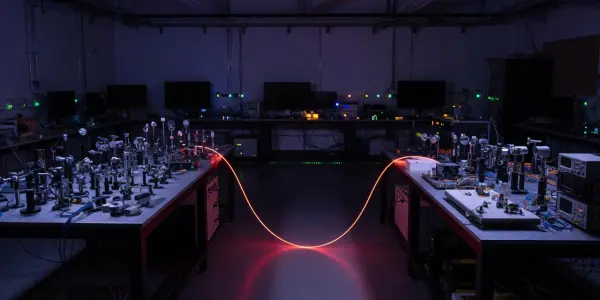A darkened laboratory contains two workbenches with optical equipment, illuminated by a bright red laser beam across the floor.