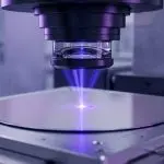 A futuristic, dark laboratory setting features a precision instrument with a purple laser focused on a circular wafer.