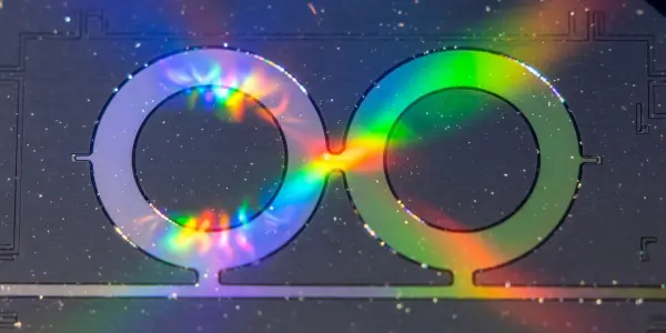 An iridescent infinity symbol glows against a dark, speckled background with circuit-like patterns, featuring blues, greens, and purples.
