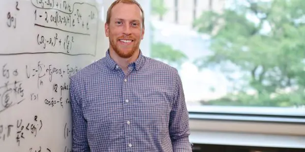 Man in button-down shirt next to whiteboard with physics equations
