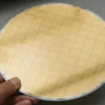A hand holds a circular silicon wafer with a grid pattern, against a dark and light grey background.