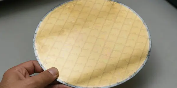 A hand holds a circular silicon wafer with a grid pattern, against a dark and light grey background.
