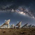 A wide-angle night view reveals numerous radio telescopes against a vibrant, star-filled sky, with earthy tones below