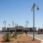 Security cameras on poles guard the gated entrance of a fenced desert facility
