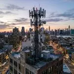 A tall communications tower rises above a dense cityscape at dusk, featuring warm golden and cool blue tones