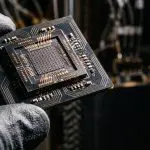 Gloved fingers holding a dark chip with a gridded core and gold wire bonds