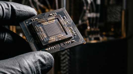 Gloved fingers holding a dark chip with a gridded core and gold wire bonds