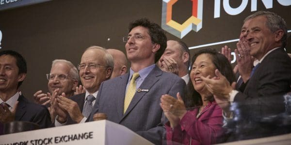 Several people are celebrating with their hands raised in the air, in a brightly lit stock exchange setting.