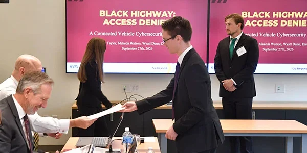Presenter hands a paper to a seated attendee as screens display a connected vehicle cybersecurity presentation