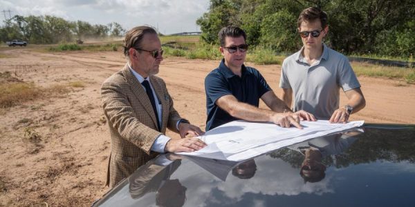 Three men in sunglasses review a large site plan spread across the hood of a car on cleared land at the Occam Foundry ca